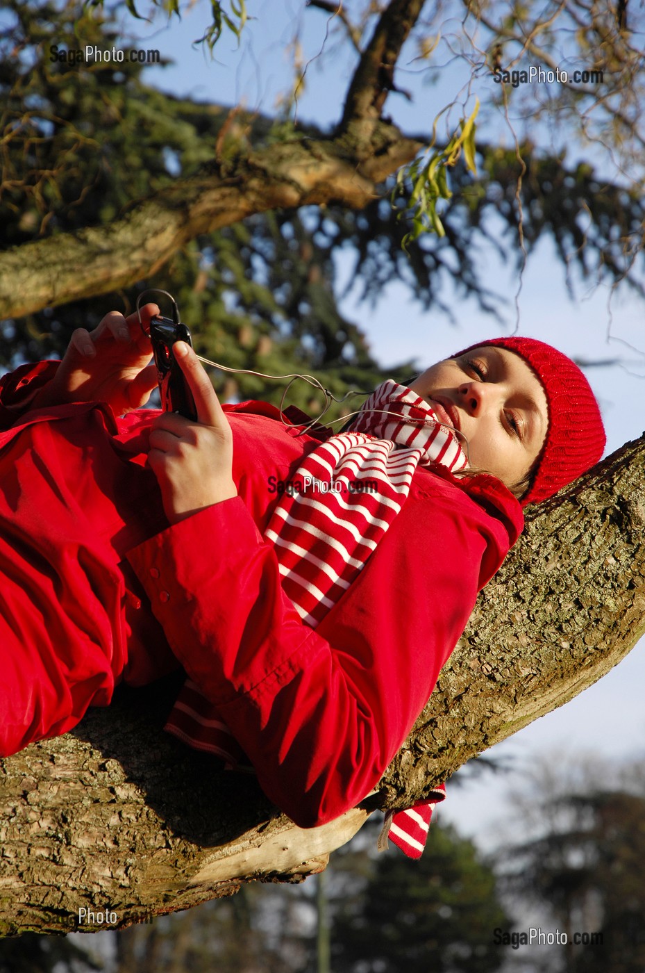 NATURE ET TECHNOLOGIE. JEUNE FEMME ECOUTANT DE LA MUSIQUE AVEC SON TELEPHONE PORTABLE, SUR UNE BRANCHE D'ARBRE, EN FORET 