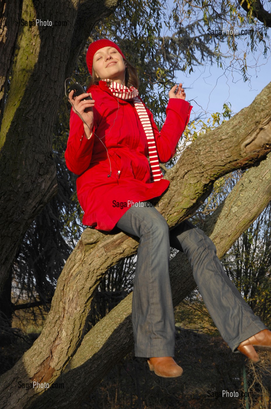 NATURE ET TECHNOLOGIE. JEUNE FEMME ECOUTANT DE LA MUSIQUE AVEC SON TELEPHONE PORTABLE, SUR UNE BRANCHE D'ARBRE, EN FORET 
