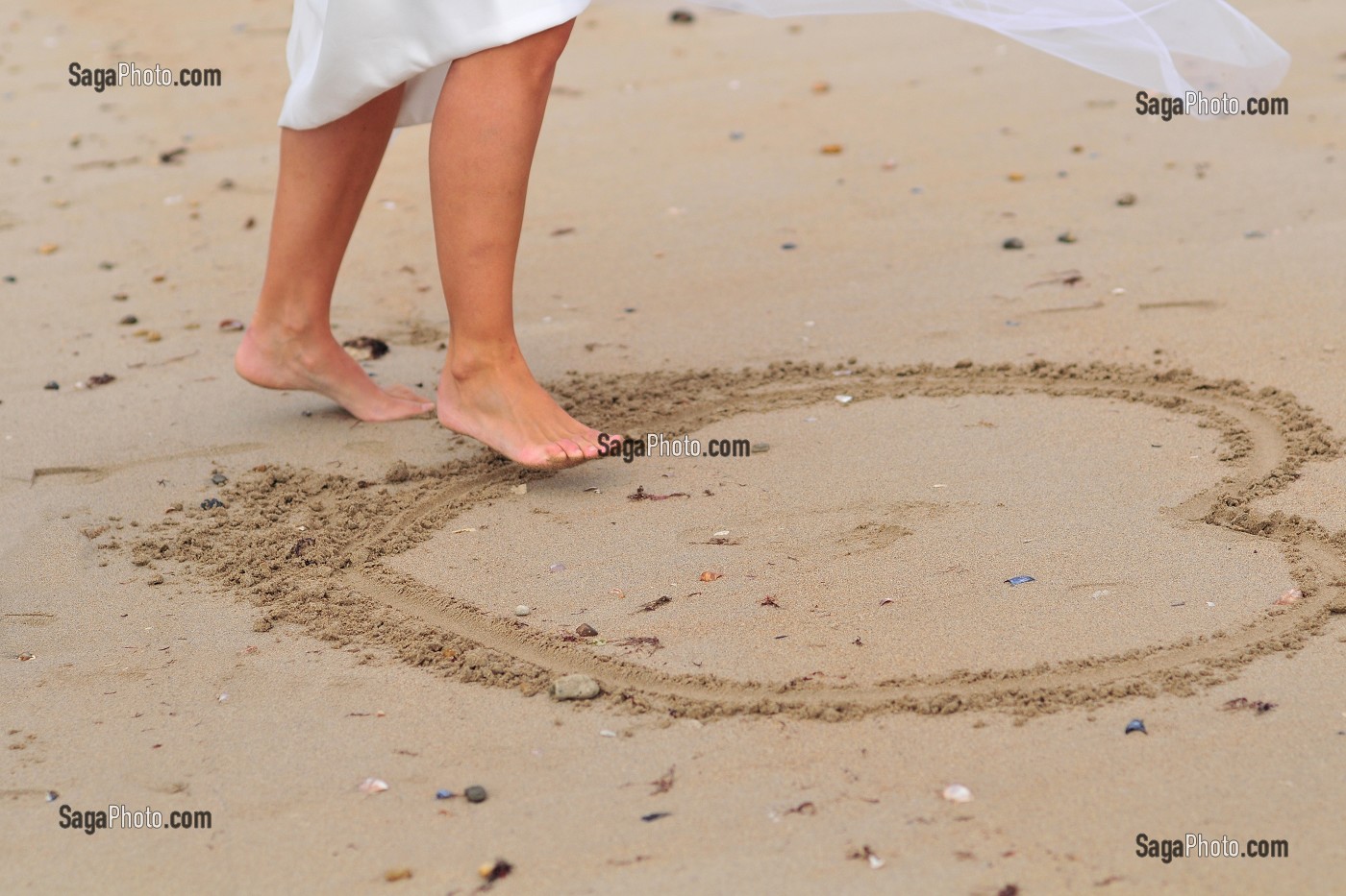 JEUNE FEMME EN ROBE DE MARIEE DESSINANT UN COEUR SUR LE SABLE AVEC SON PIED 