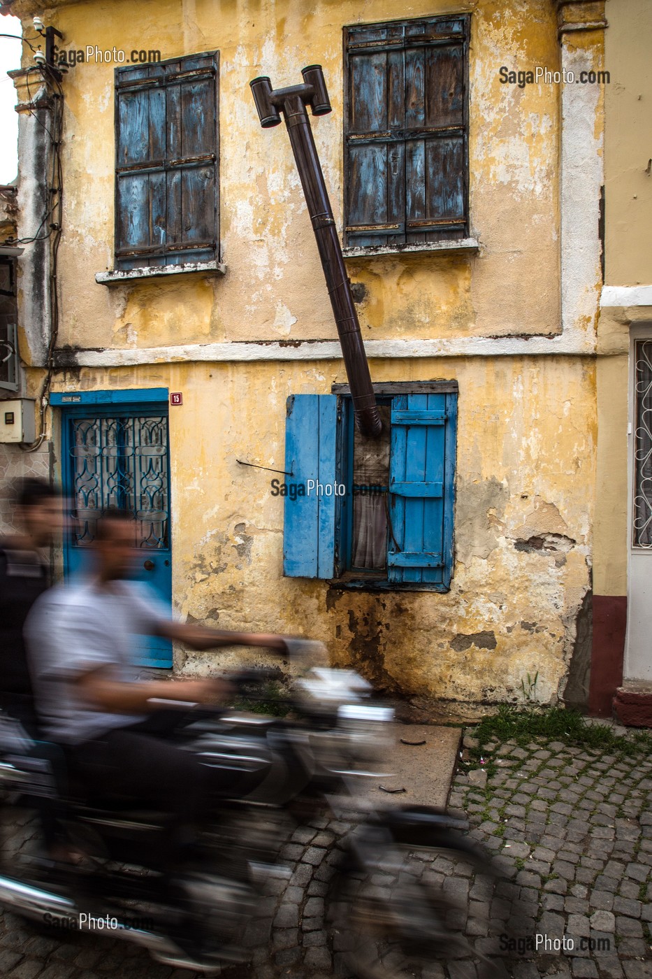 SCENE DE RUE AVEC MOTO DEVANT UNE MAISON JAUNE AVEC UNE CHEMINEE QUI SORT PAR LA FENETRE, RUE MERKEZ HASTANE, VILLE D'AYVALIK SUR LES BORDS DE LA MER EGEE, RIVIERA DES OLIVIERS, NORD D’IZMIR, TURQUIE 