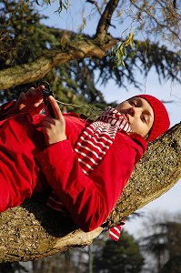 NATURE ET TECHNOLOGIE. JEUNE FEMME ECOUTANT DE LA MUSIQUE AVEC SON TELEPHONE PORTABLE, SUR UNE BRANCHE D'ARBRE, EN FORET 