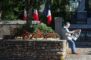 PERSONNE AGEE LISANT SON JOURNAL SUR LA PLACE DU VILLAGE, DEVANT LE MONUMENT AUX MORTS ET LES DRAPEAUX FRANCAIS, SAINT-COME D'OLT, LABEL PLUS BEAU VILLAGE DE FRANCE, AVEYRON (12), FRANCE 
