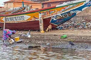 FEMME SENEGALAISE SUR LE BORD DU FLEUVE DEVANT UNE PIROGUE TRADITIONNEL ET TAS D'ORDURES DE DECHETS PLASTIQUES, GUET NDAR, QUARTIER DU VILLAGE DES PECHEURS, SAINT-LOUIS-DU-SENEGAL, SENEGAL 