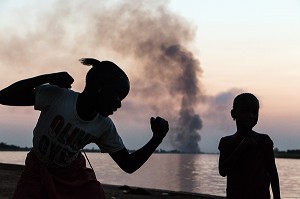 JEU D'ENFANTS DEVANT LE FLEUVE AVEC LES FEU DE CANNE A SUCRE SUR FOND DE COUCHER DE SOLEIL, DAGANA, SENEGAL, AFRIQUE DE L'OUEST 
